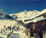 Vue de côté du restaurant d'altitude, bâtiment en bois, terrasse extérieure avec chaises longues le long des pistes enneigées, panneau 