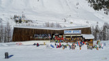 Vue de face du restaurant d'altitude, bâtiment en bois, terrasse extérieure avec chaises longues le long des pistes enneigées, panneau 