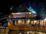 Vue nocturne du restaurant, chalet en bois et pierre de 3 étages avec terrasse extérieure au RDC, tables et chaises. Banderole 