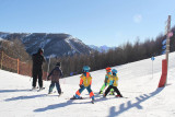 Enfants et adulte descendant une piste de ski enneigée, forêt et montagnes en arrière-plan