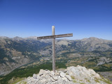 A wooden cross situated at the summit of Rochecline Mountain, on a pile of stones. A completely unobstructed view of the village of Allos and the mountains below