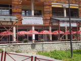 Restaurant frontage in summer with outdoor terrace furnished with tables, chairs, and red parasols. Sign with the inscription: 
