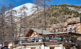 Overview of the wooden restaurant, located in front of buildings in the ski resort, atop a two-story building, with a terrace. 