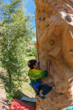 A child wearing a harness, climbing a climbing wall located in the forest above a mattress