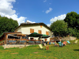 Overview of the restaurant, a two-story stone and wood building with a wooden terrace on the ground floor, tables, chairs, parasols, and sun loungers. Meadow in front of the restaurant with sun loungers.