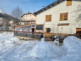 Overview of the restaurant in winter: a two-story stone and wood building with a wooden terrace on the ground floor, tables, and chairs. Snow-covered meadow in front of the restaurant. Wooden sign reading 
