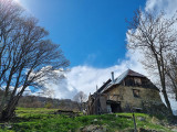 Stone house with a corrugated iron roof situated in a meadow surrounded by a few trees. Forest in the background.