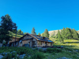 Vue du refuge situé au bord du Lac d'Allos, maison en bois comportant une terrasse avec des tables et chaises, dans un cadre de végétation et de conifères