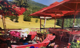 The restaurant's outdoor terrace in summer, featuring plastic tables and chairs, large parasols, and a view of a field and the mountains.