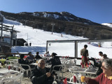 The restaurant's outdoor terrace in winter, featuring plastic tables and chairs, views of the snowy slopes and mountains, and a nearby chairlift.