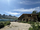 Side view of the wooden refuge, terrace with tables and chairs. Lake Allos visible nearby, with the Tours du Lac in the background.
