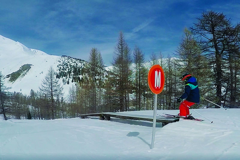 Piste ludique de ski entourée de conifères, enfant skieur s'apprêtant à glisser sur une rampe
