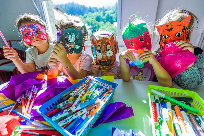 Children with handmade animal and fruit paper masks, Giotto crayons on the table