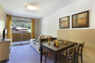 Séjour lumineux d'appartement avec canapé, meuble TV, TV, tabl à manger, porte-fenêtre donnant sur un balcon, vue sur la station