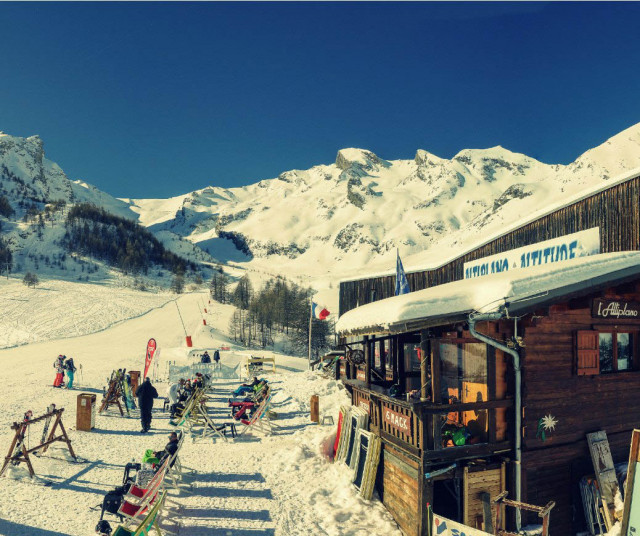 Vue de côté du restaurant d'altitude, bâtiment en bois, terrasse extérieure avec chaises longues le long des pistes enneigées, panneau 