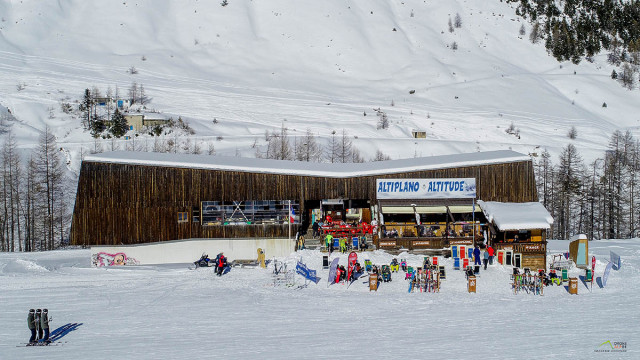 Vue de face du restaurant d'altitude, bâtiment en bois, terrasse extérieure avec chaises longues le long des pistes enneigées, panneau 