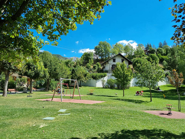 Aire de jeux pour enfants située au milieu d'une prairie verdoyante, balançoire, bancs et chaise à bascule
