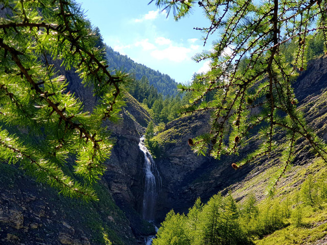 Cascade visible en fond au milieu d'une formation rocheuse entourée par la végétation. Montagnes boisées en arrière-plan