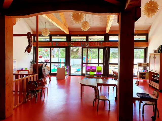 Nursery reception hall, colorful walls, tables, chairs, children's toys, outdoor courtyard accessible through French doors. Sign reads 