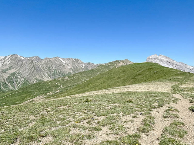View of the e-MTB route at the summit of the Col d'Allos, a high-altitude trail across a mountain meadow with mountains in the background.