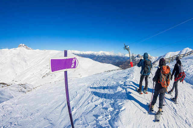 3 randonneurs en raquette au sommet d'un sentier de randonnée se situant près d'un téléski visible, grande surfaces enneigées et montagnes