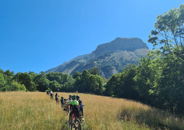 VTTistes progressant sur le plateau de Montgros, prairie en été entourée d'arbres, montagne de Montgros en arrière-plan