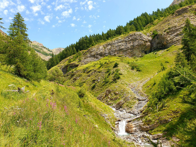 A stream winding through a mountain valley, with the Cascade du Cimet hiking trail visible in the middle, and forest and mountains in the background