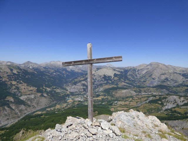 A wooden cross situated at the summit of Rochecline Mountain, on a pile of stones. A completely unobstructed view of the village of Allos and the mountains below