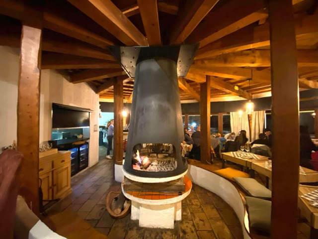 The restaurant's interior dining room, centered around a large fireplace, offers a plush atmosphere with tables and chairs, benches, and a TV.