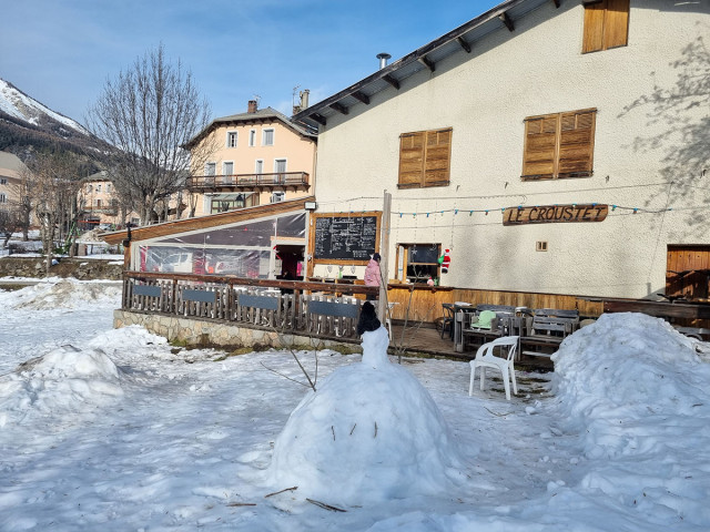 Overview of the restaurant in winter: a two-story stone and wood building with a wooden terrace on the ground floor, tables, and chairs. Snow-covered meadow in front of the restaurant. Wooden sign reading 