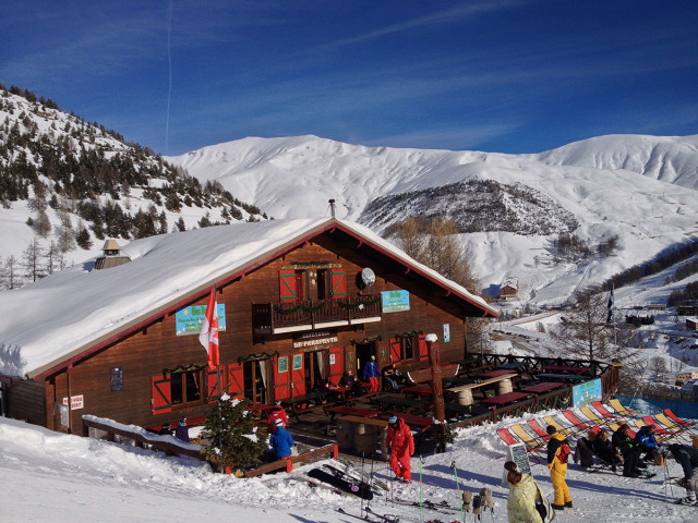 Vue du restaurant, chalet en bois, avec sa terrasse, tables et chaises, chaises lonuges. Montagnes enneigées tout autour et en arrière-plan - © Le Parapente B Ollivier Vue du restaurant, chalet en bois, avec sa terrasse, tables et chaises, chaises lonuges. Montagnes enneigées tout autour et en arrière-plan