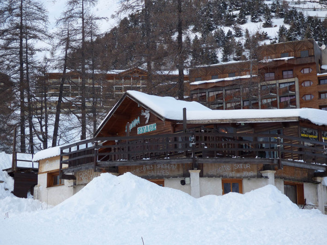 Overview of the wooden restaurant during winter, located in front of buildings in the ski resort, atop a two-story building, with a terrace. 