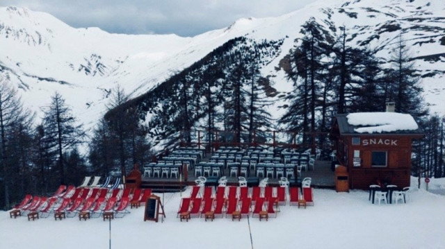 Vue du restaurant de face, chalet et terrasse en bois au milieu des pistes et de la forêt, tables et chaises, chaises longues. Montagnes en arrière-plan