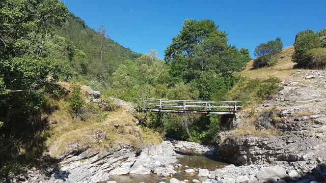 Baumelle Bridge, a footbridge situated over a stream winding between rocks and trees. Surrounding path: rocky, with vegetation, groves, and trees