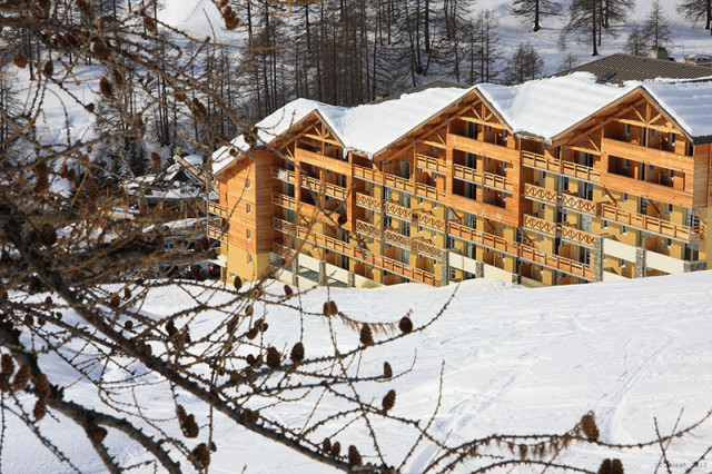 Vue de côté de la résidence, bâtiment sur plusieurs étages, neige sur les toits, forêt en arrière-plan - © Les cimes du Val d'Allos Vue de côté de la résidence, bâtiment sur plusieurs étages, neige sur les toits, forêt en arrière-plan