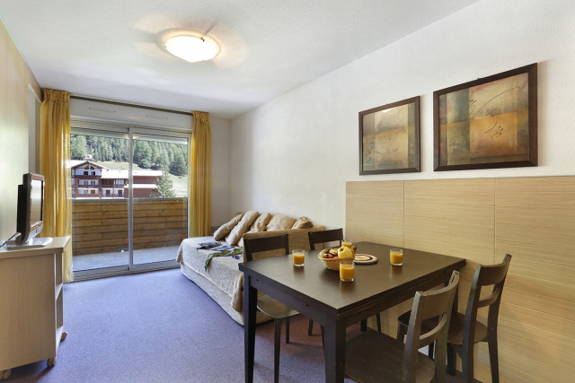 Séjour lumineux d'appartement avec canapé, meuble TV, TV, tabl à manger, porte-fenêtre donnant sur un balcon, vue sur la station
