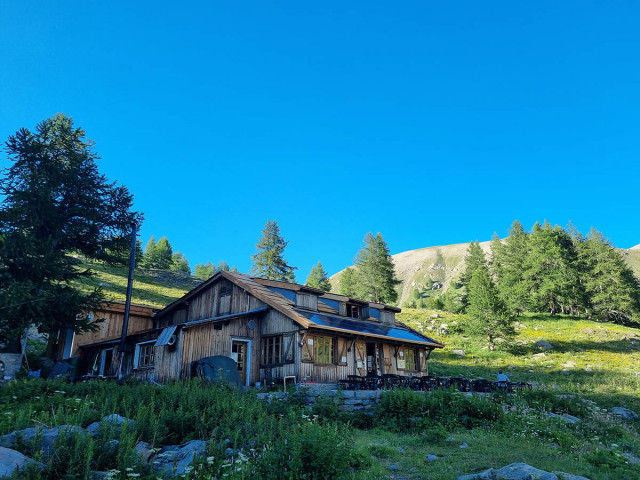 Vue du refuge situé au bord du Lac d'Allos, maison en bois comportant une terrasse avec des tables et chaises, dans un cadre de végétation et de conifères