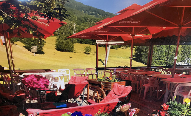 The restaurant's outdoor terrace in summer, featuring plastic tables and chairs, large parasols, and a view of a field and the mountains.