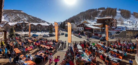 Terrasse de la brasserie avec une grande foule, tables et chaises, située à côté de la place centrale de la Foux, vision sur la station