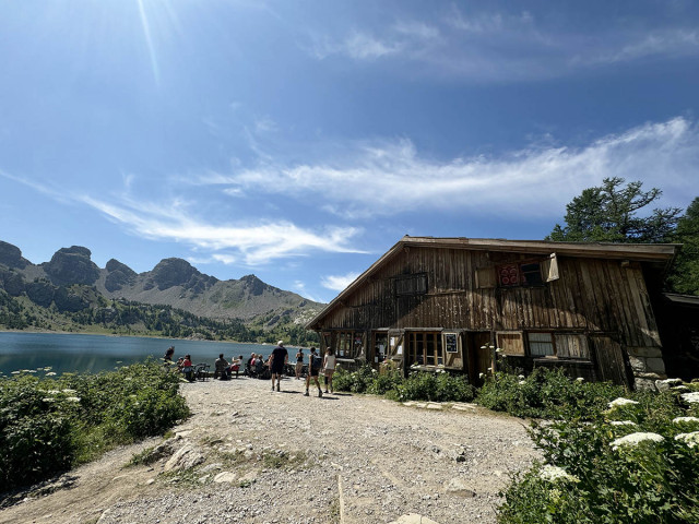 Side view of the wooden refuge, terrace with tables and chairs. Lake Allos visible nearby, with the Tours du Lac in the background. - © B Courbon Side view of the wooden refuge, terrace with tables and chairs. Lake Allos visible nearby, with the Tours du Lac in the background.