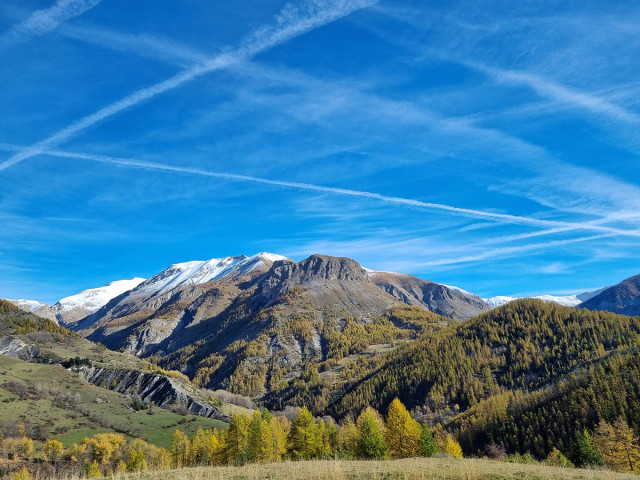 An unobstructed view of a colorful, wooded mountain valley. The mountain peaks in the background are snow-capped.