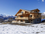 Vue de côté d'un grand chalet isolé de 3 étages en bois et pierre, grandes terrasses en bois et baies vitrées, paysage montagnard