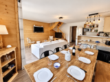 Grande pièce de vie chaleureuse, avec une décoration en bois style chalet de montagne. Une grande table en bois dressée fait face au salon avec canapé et télévision. La cuisine ouverte et escalier en bois au fond de la pièce.