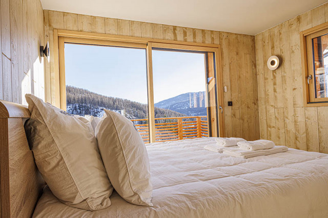 Chambre à coucher lumineuse lit deux places, ameublement bois, porte-fenêtre donnant sur un balcon en bois Chambre à coucher lumineuse lit deux places, ameublement bois, porte-fenêtre donnant sur un balcon en bois