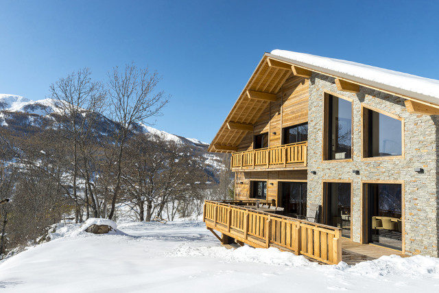 Vue de côté d'un grand chalet en bois et pierre sur deux étages, nombreuses fenêtres et large terrasse, balcon au 1er étage. Paysage enneigé. - © M Desvignes Vue de côté d'un grand chalet en bois et pierre sur deux étages, nombreuses fenêtres et large terrasse, balcon au 1er étage. Paysage enneigé.