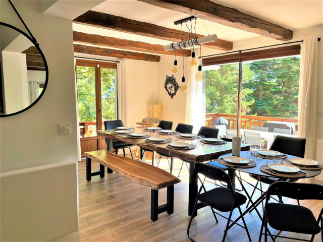 Séjour lumineux avec table à manger, ameublement bois, poutres au plafond, portes-fenêtres donnant sur une terrasse en bois
