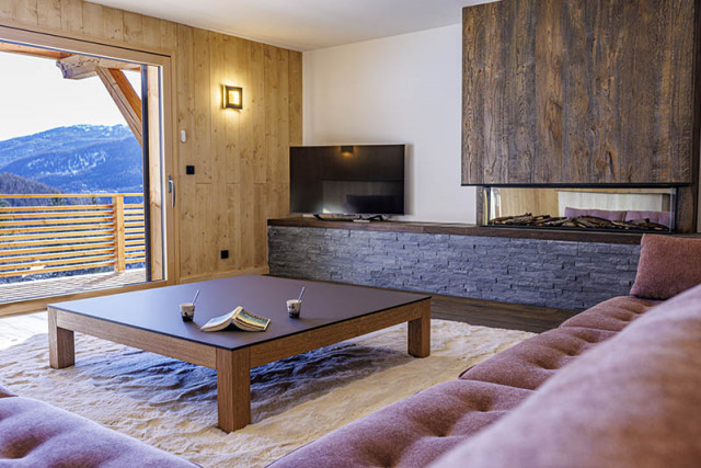 Large, bright living room with coffee table, sofa, television, wooden and stone furnishings, French windows opening onto a wooden balcony with snowy views.