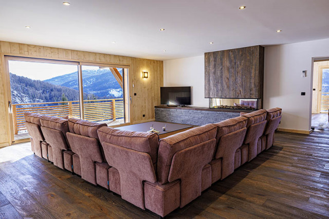 Large, bright living room with coffee table, sofa, television, wooden and stone furnishings, French windows opening onto a wooden balcony with snowy views.