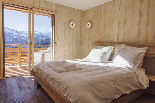 Spacious and bright bedroom with double bed, wooden furniture and parquet flooring, French windows opening onto a wooden balcony and a snow-covered landscape.