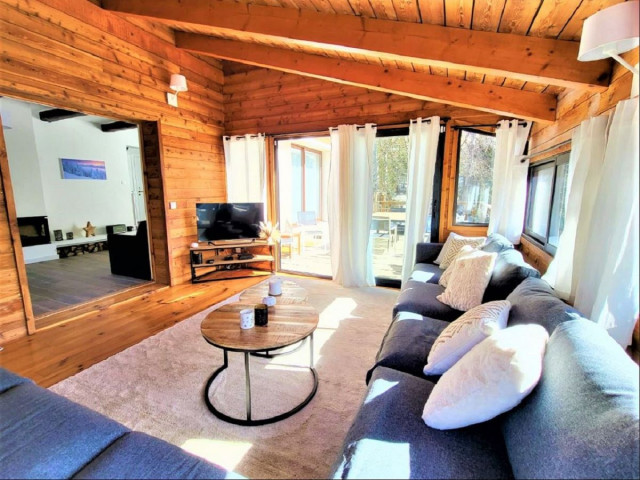 Séjour lumineux dans une véranda en bois, avec canapé, tapis, table basse, TV, porte-fenêtre, jonction avec un salon intérieur carrelé